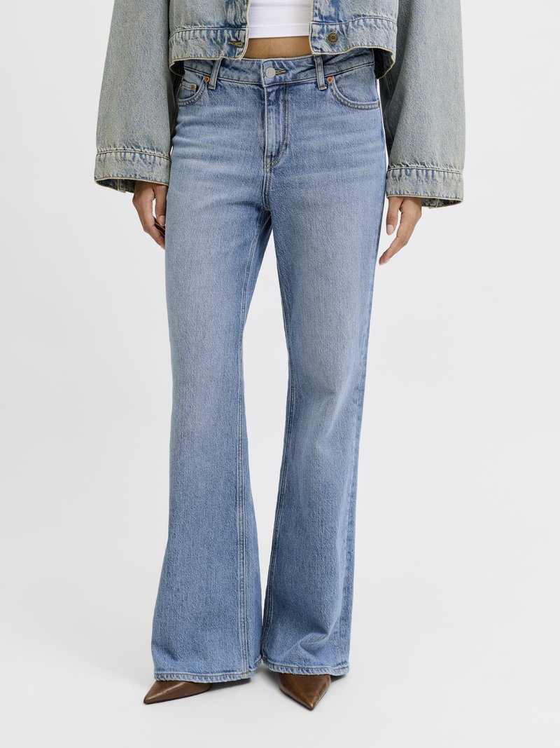 Lyseblå buks med høj talje og udvidede ben, båret med en kort denimjakke, hvid top og brune spidse sko, vist fra midt på torso til fødder.