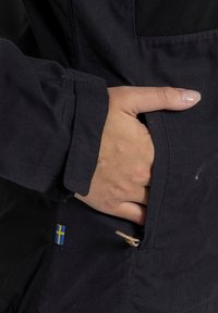 Black outerwear jacket featuring a side pocket, a textured finish, and a small Swedish flag accent near the bottom hem.