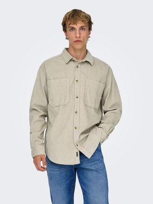 Junger Mann trägt ein beiges Hemd mit Knöpfen und zwei Brusttaschen und blaue Jeans, steht mit einer Hand in der Hosentasche.