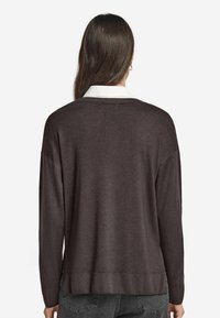 Dunkelbrauner, weicher Strickpullover mit entspanntem Schnitt, geripptem Saum und überschnittenen Schultern, über einem weißen Hemd mit Kragen getragen.