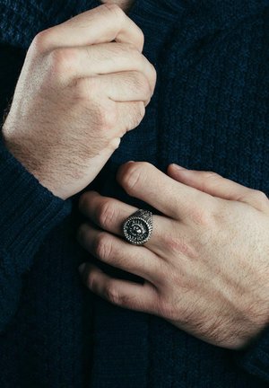 Struktureran temno modra pulover, roke zložene. Srebrn spiralen prstan ima zapletene vzorce in osrednji kamen.