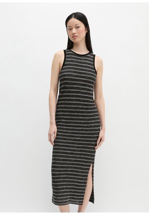 Vrouw draagt een mouwloze midi-jurk met zwart-witte horizontale strepen en een zijsplit, staand tegen een effen lichte achtergrond.
