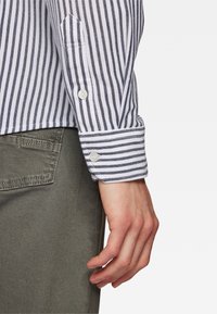 Camisa a rayas con líneas verticales azules y blancas, puños abotonados con dos botones, combinada con pantalones verde oliva. Detalle de tejido texturizado visible.