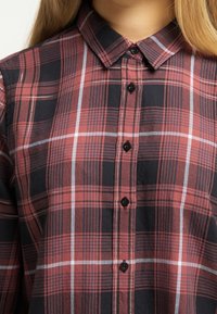 Röd och svart rutig skjorta med knappar, med spridd krage, som har svarta knappar och vita accentränder i mönstret. Mjuk tygstruktur.