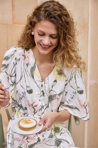 Femme aux cheveux bouclés souriante tenant une assiette avec un dessert rond surmonté de meringue toastée, vêtue d'une robe fleurie.