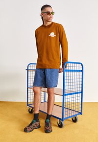 Brun långärmad skjorta med logotyp framtill, blå jeansshorts och bruna vandringsskor. En blå metallvagn står i närheten.