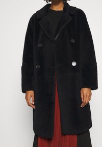 Svart dubbelknäppt kappa gjord av plyschmaterial med krage, stora knappar och ett logopatch på framsidan, med breda ärmar.