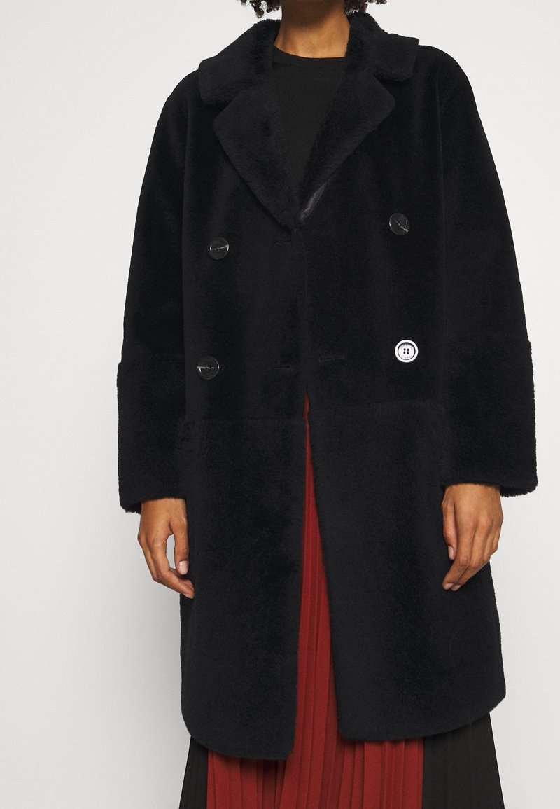 Svart dubbelknäppt kappa gjord av plyschmaterial med krage, stora knappar och ett logopatch på framsidan, med breda ärmar.