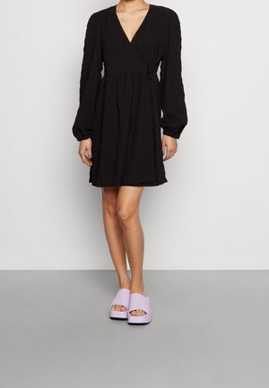 Robe noire portefeuille à manches longues et en tissu texturé, associée à des sandales à plateforme ouvertes violet lavande. Modèle longue longueur.