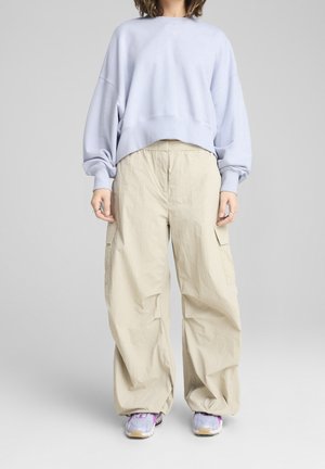Personne portant un sweat oversize violet clair, un pantalon cargo beige à jambes larges et des baskets blanches avec des accents violets.