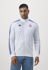 Hvit zip-jakke med blå striper på ermene, med Real Madrid-logo på brystet. Laget av glatt stoff, tettsittende design, enkel krage.