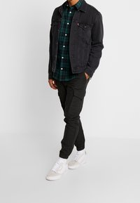 Schwarze Jeansjacke über einem grün-karierten Hemd, kombiniert mit dunklen Cargo-Hosen und weißen Sneakers. Einfaches Design mit sichtbaren Nähten.