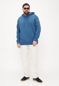 Felpa blu in cotone con tasca frontale, cappuccio con lacci e emblema del logo. Abbinata a pantaloni crema e scarpe nere. Texture morbida.
