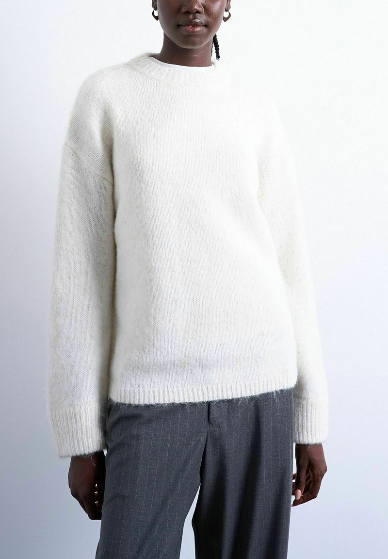 Personne noire portant un pull en tricot blanc duveteux et un pantalon gris à rayures fines sur un fond clair uni.