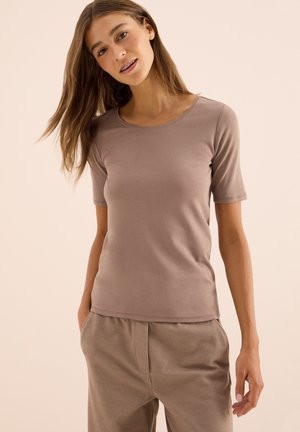 Jeune femme avec de longs cheveux bruns, portant un T-shirt taupe à manches courtes et un pantalon assorti, se tenant avec les mains dans les poches devant un fond beige.