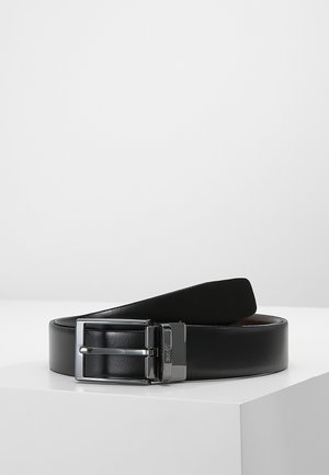 Cintura in pelle nera con fibbia rettangolare argentata lucida, arrotolata e appoggiata su una superficie bianca su uno sfondo chiaro e uniforme.