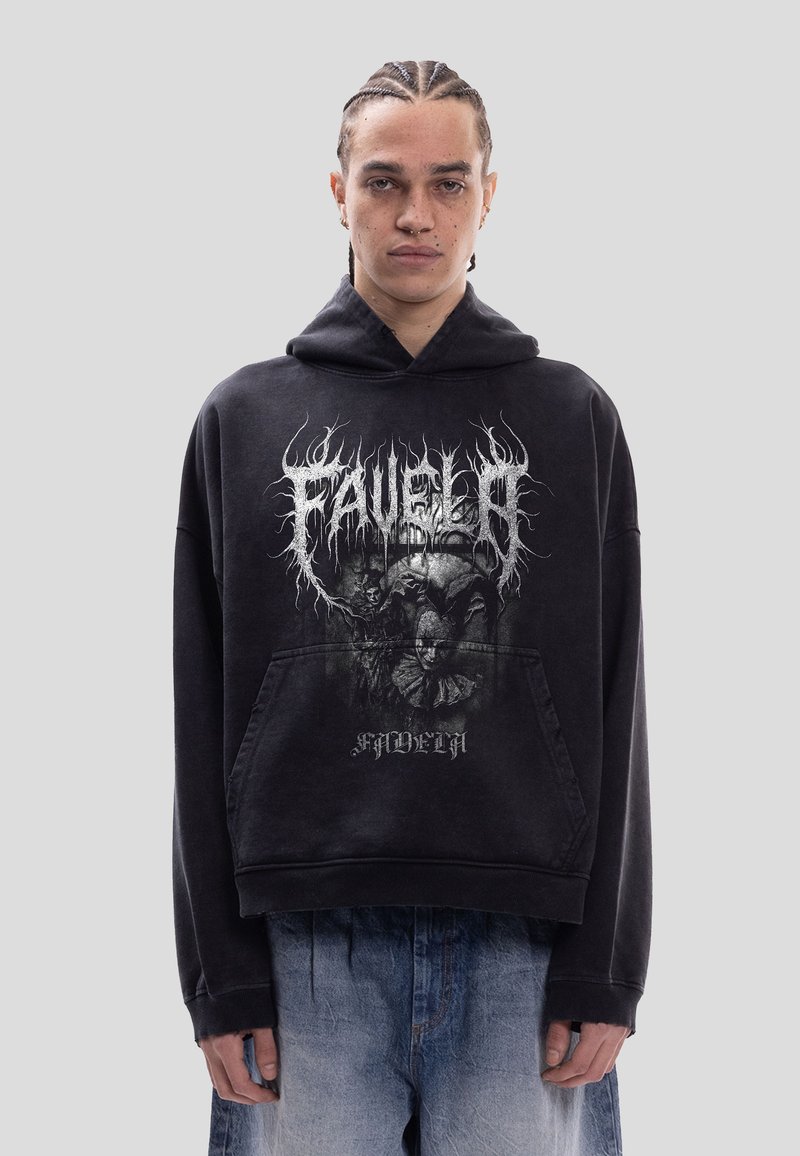 Personne aux cheveux tressés portant un sweat à capuche noir avec un texte gothique et des graphismes de clown, associée à un jean bleu ample.
