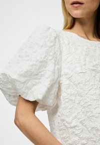 Camicia bianca con texture, maniche corte a sbuffo, caratterizzata da un motivo increspato su tutta la superficie del tessuto. Collo rotondo, design semplice.