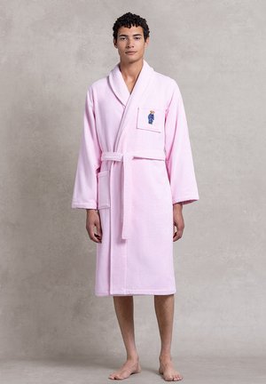 Robe en éponge rose avec ceinture, présentant un patch d'un ours en vêtements bleus sur la poche poitrine. Coupe ample avec des manches longues.