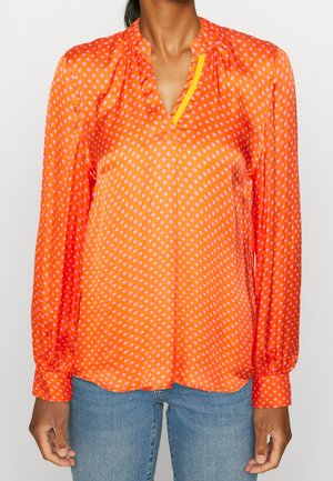 Woman wearing an orange long-sleeve blouse with white polka dots and a yellow trim, paired with blue jeans.