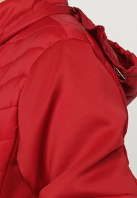 Chaqueta acolchada roja con costuras acolchadas en la zona del hombro y mangas de tela suave, mostrando una vista en primer plano de la parte superior del brazo y el hombro.