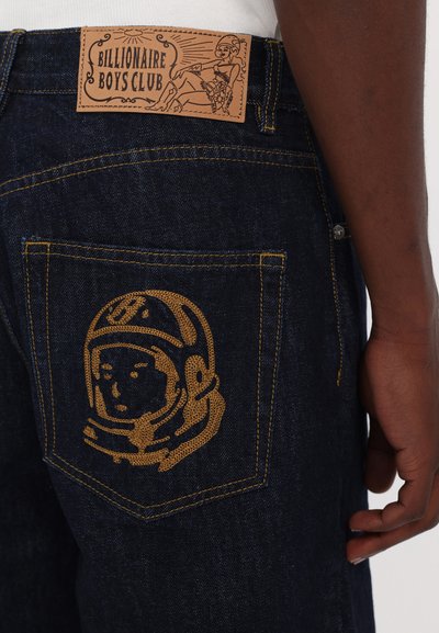 Džíny z denimu s tmavým umytím, so žltým stehom a grafikou astronauta na zadnom vrecku. Kožená nášivka zobrazuje "Billionaire Boys Club."