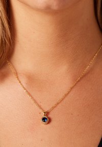 Gouden ketting met een ronde blauwe hanger omringd door heldere stenen, rustend op de huid. De ketting is fijn met een schakeldesign.