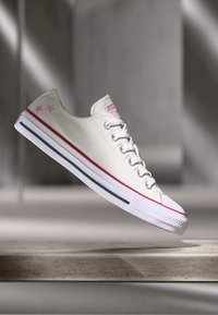 Canvas low-top sneaker in off-white with pink star accents, red and blue striped sole, and white rubber toe cap. Features six eyelets and laces.