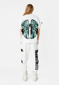 Camiseta blanca con un gran diseño de alas de ángel multicolores en la parte posterior, combinada con pantalones cargo blancos que tienen detalles en negro y texturas.