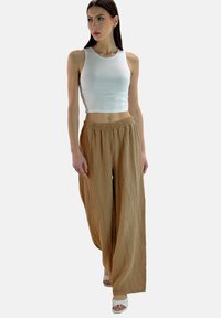 Beige, gestructureerde broek met wijde pijpen, een relaxte pasvorm en een elastische tailleband, gecombineerd met een getailleerd wit tanktop en lichte sandalen.