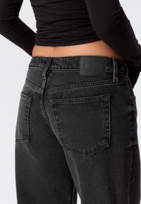 Personne portant un jean en denim noir taille haute avec des poches arrière visibles et un patch en cuir noir portant l'inscription "WEEKDAY".