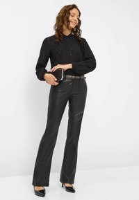 Blouse noire texturée à manches longues bouffantes, assortie à un pantalon en cuir noir ajusté et des talons aiguilles noirs. Tient une petite pochette.