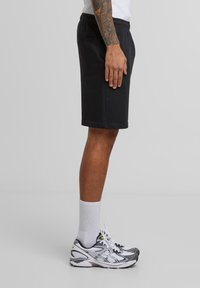 Male wearing black knee-length shorts, white crew socks, and white-gray running shoes, standing sideways against a plain background.