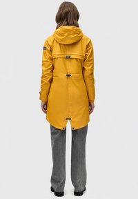 Waterdichte gele regenjas met capuchon, verstelbare taille, achterste ventilatie en drukknoopsluitingen. Gecombineerd met grijze wijde broek en zwarte schoenen.