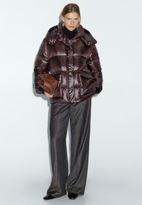 Donna che indossa un giubbotto lucido marrone scuro, pantaloni larghi grigi, scarpe nere, e tiene in mano una pochette di pelle marrone su uno sfondo semplice.