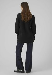 Maglione nero a maglia con texture, maniche lunghe, abbinato a jeans a gamba larga scuri e calzature nere, visto da dietro su uno sfondo neutro.