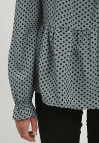 Camicia grigia a pois neri, design peplum, maniche lunghe con polsini arricciati, realizzata in tessuto morbido. Indossata su pantaloni neri.