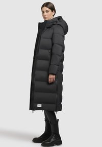 Schwarze Pufferjacke mit langem, gesteppte Design, ausgestattet mit Kapuze und Reißverschluss vorn. Begleitet von schwarzen Knöchelstiefeln und figurbetonten Leggings.