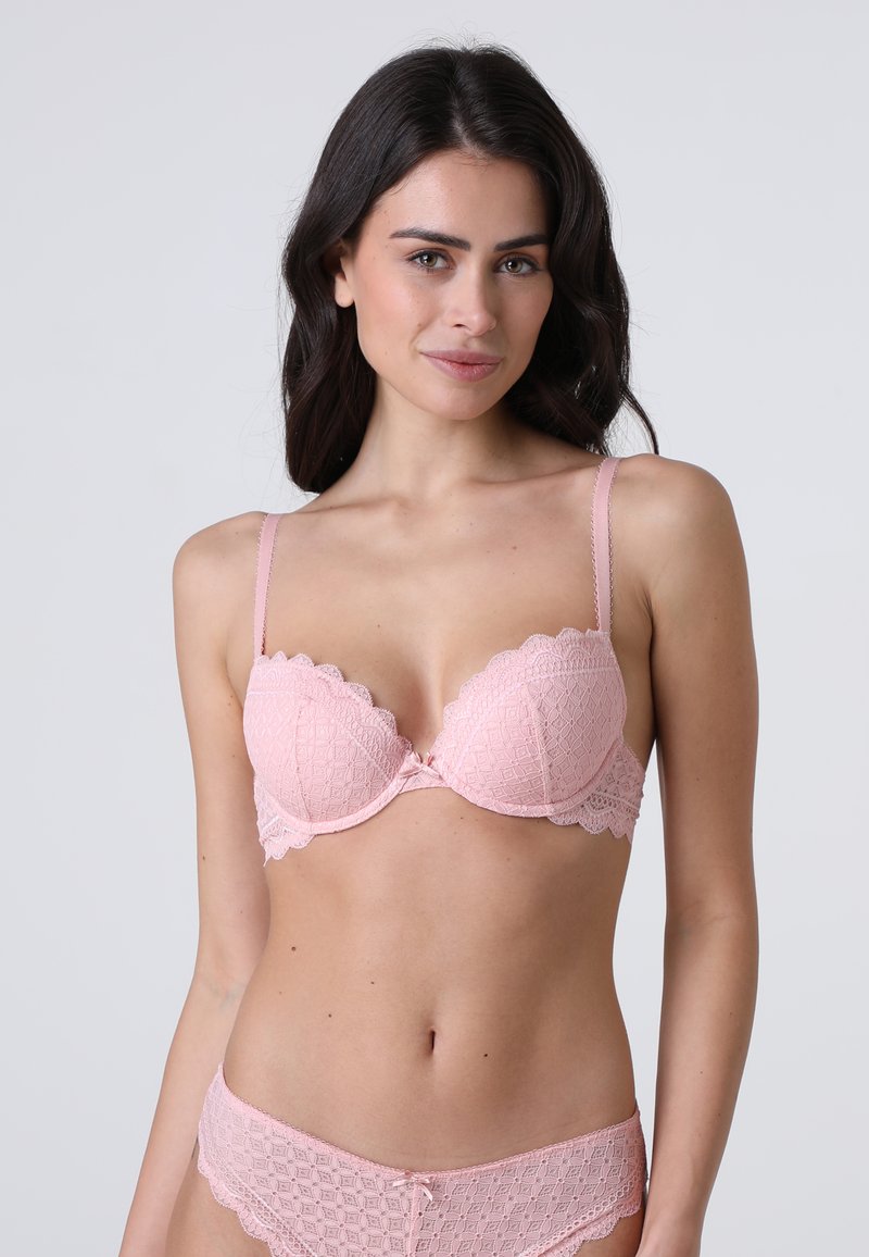 Donna con lunghi capelli scuri che indossa un completo coordinato di reggiseno e slip in pizzo rosa chiaro, in piedi contro uno sfondo grigio chiaro uniforme.