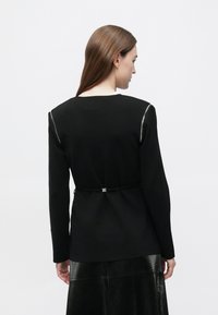 Žena s dlhými hnedými vlasmi, oblečená v čiernom top s dlhým rukávom a zipsami na ramienkach a v čiernej štruktúrovanej sukni, otočená chrbtom.