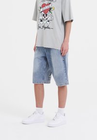 Camiseta gráfica de color gris claro con diseño de calavera y corazón, combinada con pantalones cortos de mezclilla azul y zapatillas blancas. Ajuste casual y relajado.