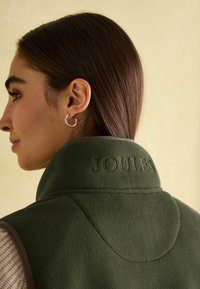 Grüne Fleece-Weste mit hohem Kragen, geprägtem "JOULES"-Logo auf dem Rücken und braunen Akzenten. Glatte Textur mit strukturiertem Schnitt.