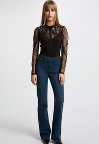 Top nero con maniche trasparenti in pizzo e pois, abbinato a jeans a vita alta blu con due tasche frontali e una leggera svasatura.