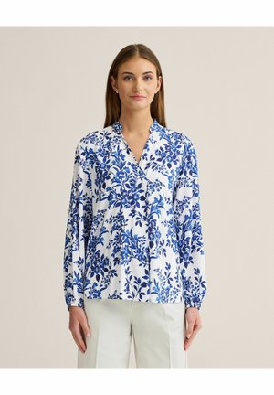 Kvinde iført en hvid bluse med blå blomsterprint og lyse bukser, stående mod en ensfarvet neutral baggrund.