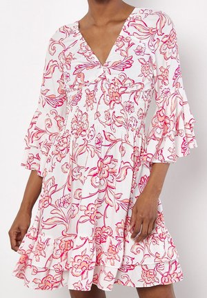 Femme portant une robe blanche avec des motifs floraux roses et orange, col en V, taille smockée et manches trois-quarts à volants ainsi que bas volanté.
