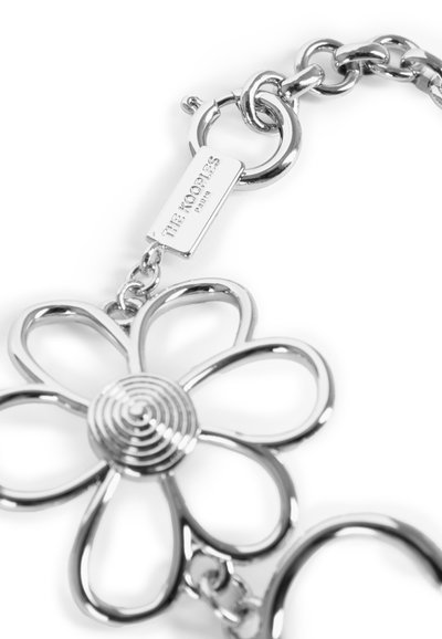 Colgante de plata en forma de flor con un centro en espiral, unido a una cadena metálica. Incluye una etiqueta con el nombre de la marca grabado. Superficies suaves y reflectantes.