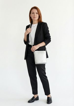 Frau in schwarzem Anzug und weißem Hemd, die eine silberne Umhängetasche hält und vor einem schlichten weißen Hintergrund steht.
