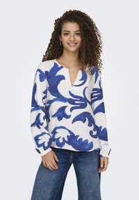 Weißes Langarmoberteil mit blauem Blumenmuster, mit V-Ausschnitt und lockerer Passform. Kombiniert mit blauen Jeans für einen Kontrast.