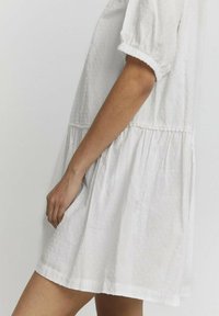 Weißes Kleid mit einem strukturierten Punktemuster, kurzen Puffärmeln und einer gerafften Taille. Knielang, mit lockerem Schnitt und weichem Stoff.
