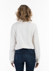 Witte blouse met lange mouwen en een kraaghals, zachte textuur en een relaxte pasvorm, gecombineerd met donkerblauwe strakke jeans met achterzakken.